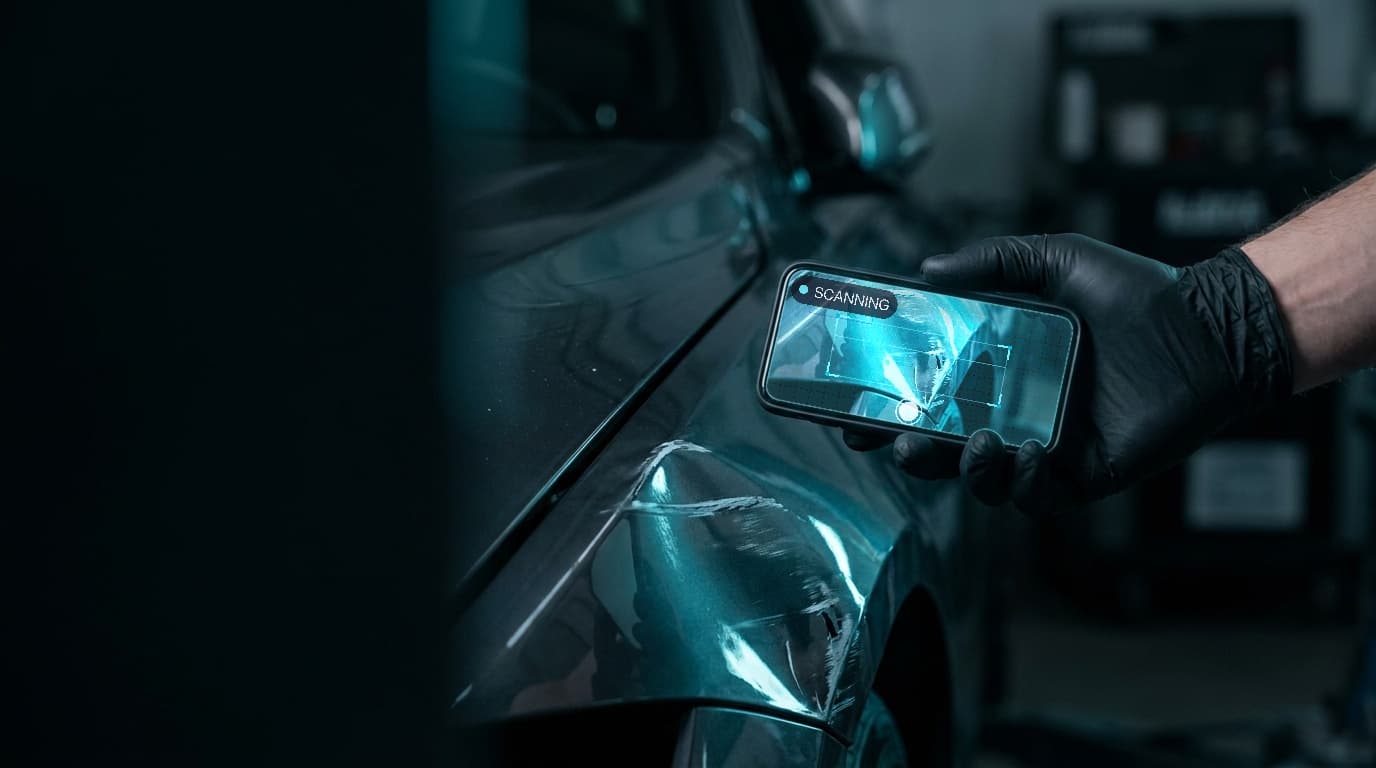 A gloved technician holds a phone up to a creased fender, the damage framed on screen by the scanner